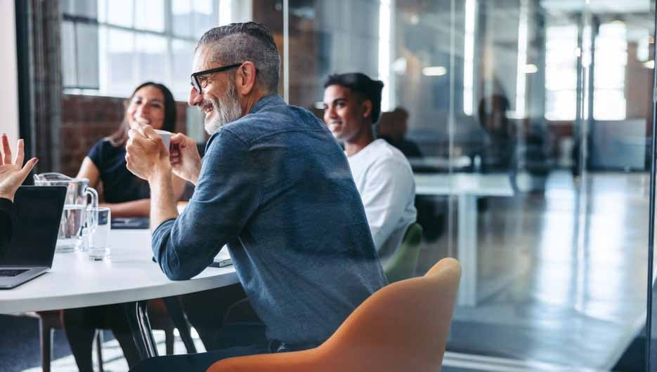 Happy mature businessman attending a meeting with his colleagues in an office. Experienced businessman smiling cheerfully while sitting with his team in a meeting room. Businesspeople working together.